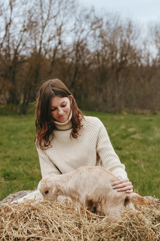 A woman in a cream knit sweater sits on straw in a field, gently stroking a goat kid, capturing a calm, affectionate farm lifestyle moment outdoors, with natural light and rural peace.