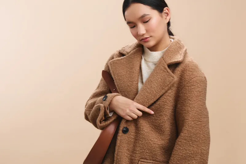 asian woman wearing a warm camel coat stands against a soft beige backdrop, looking down with a calm expression, conveying quiet confidence and contemporary style.