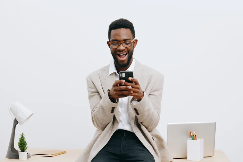 Smiling man using mobile phone sitting at desk in modern office environment, wearing smart casual attire, glasses, happy expression, freelance or business casual concept, isolated on plain white backg