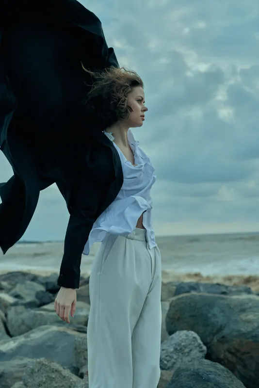 Wind sweeps through an outdoor coastal scene as a stylish woman stands among rocks, a flowing coat billowing behind her, capturing movement, mood, and fashion against the sea backdrop.