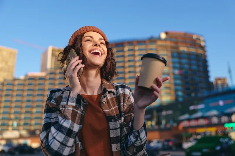 woman with coffee and phone laughing with a bright smile in an urban street scene, candid authenticity and warm golden hour glow, modern mindful living and emotional storytelling vibe.