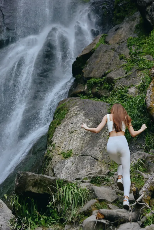 Woman hiking near waterfall wearing white activewear climbs rocky terrain surrounded by lush green foliage in misty nature setting, showcasing adventure and fitness.