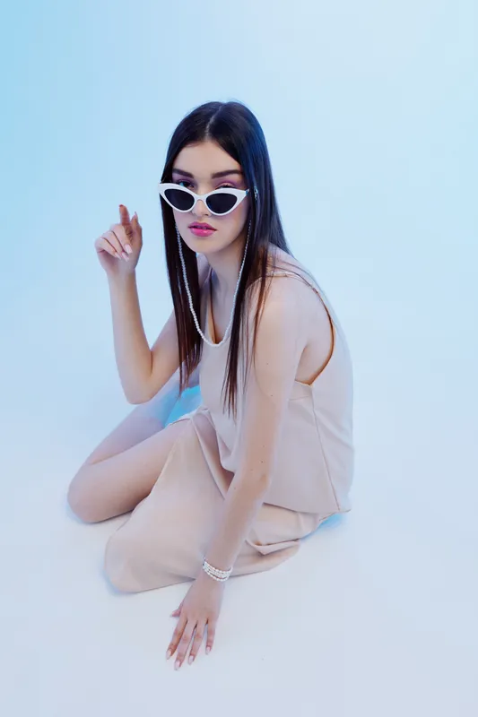 Young woman in beige outfit and white sunglasses poses sitting on the floor against a light background. Concept of beauty, fashion, style, and modern trends.