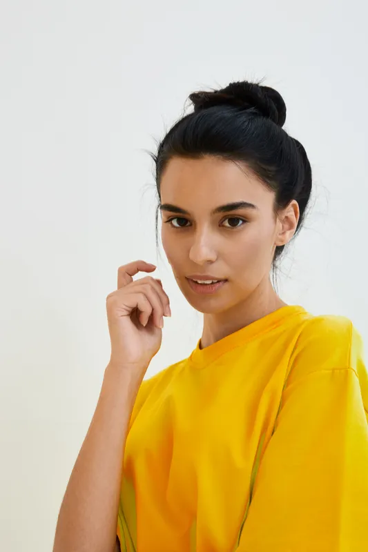 Young woman in yellow shirt with dark hair tied up, posing indoors against a white background, showcasing natural beauty and casual style.