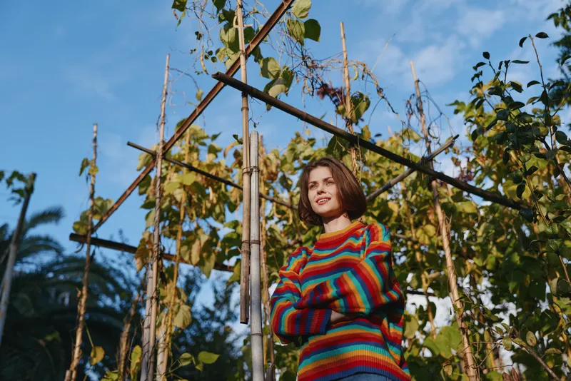 Young woman in a striped sweater with short brown hair stands by a garden trellis smiling with arms crossed in late afternoon sun, casual outfit for lifestyle and travel portrait.