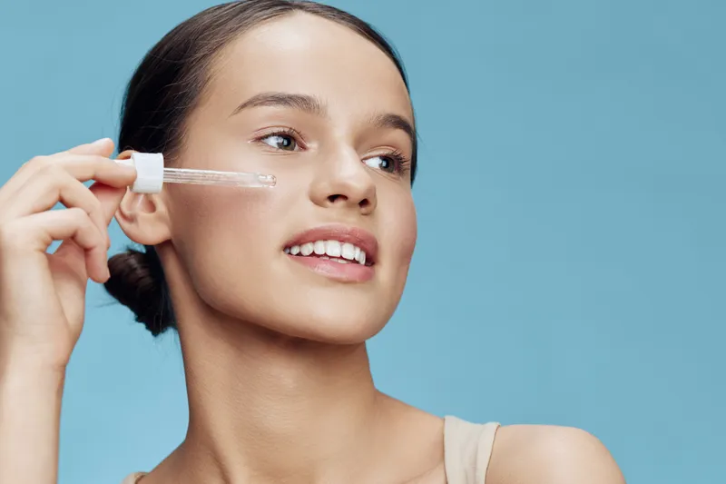 Young woman applying serum to her face, showcasing glowing skin and a joyful expression against a light blue background, representing skincare and beauty