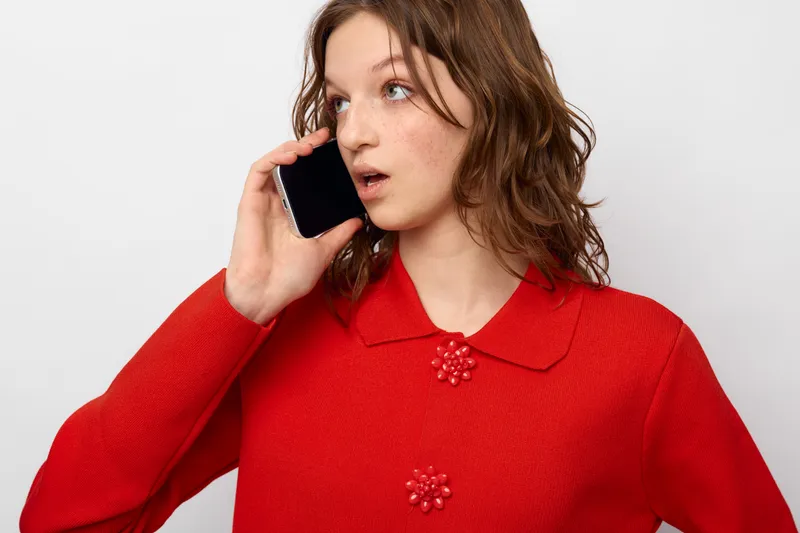 Young woman in red sweater talks on smartphone with surprised expression. Isolated on white background. Concept of communication, technology, and emotional reaction.