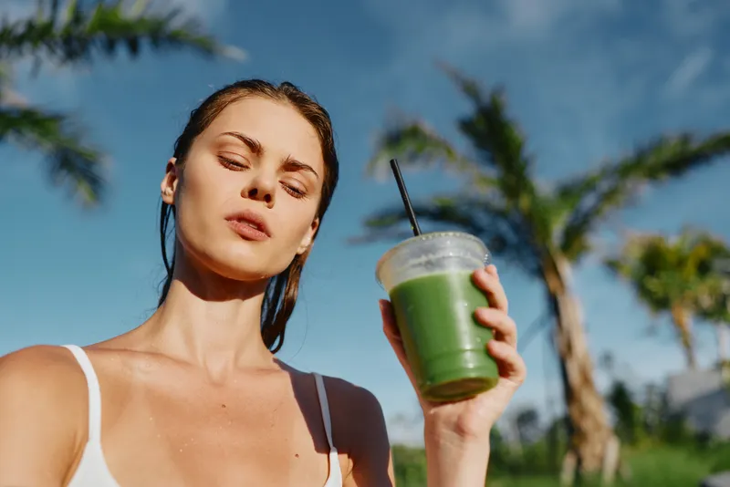 Young woman with brunette hair enjoys a green smoothie outdoors on a sunny day with palm trees in the background, expressing a relaxed and healthy lifestyle mood.