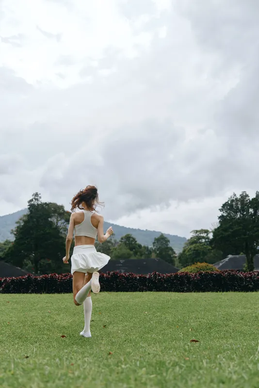 Young woman running joyfully in a grassy field, wearing a white sports outfit with a stylish skirt, capturing a sense of freedom and vitality