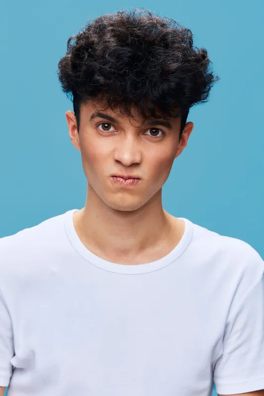 young man with curly hair wearing white t-shirt frowning with displeased expression on blue background, close-up portrait of frustrated guy