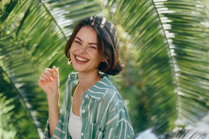 Cheerful portrait of a young woman smiling brightly amid lush tropical greenery, wearing a striped green shirt and white top, sunlit mood and relaxed vibe.