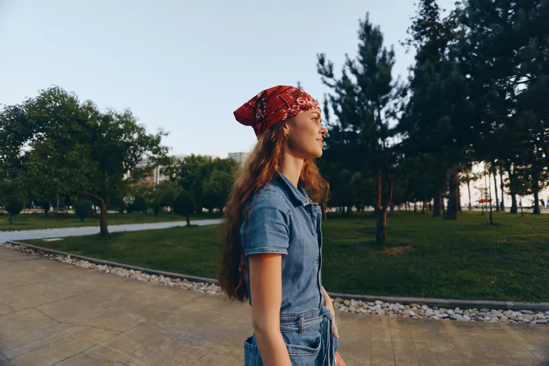 young woman wearing a red bandana and denim outfit walking outdoors in a city park during daytime with green trees and clear sky