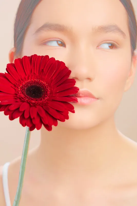 asian woman with a vivid red flower near her face, creating a soft, contemplative portrait with delicate lighting, smooth skin, and a tranquil mood ideal for beauty and lifestyle campaigns.