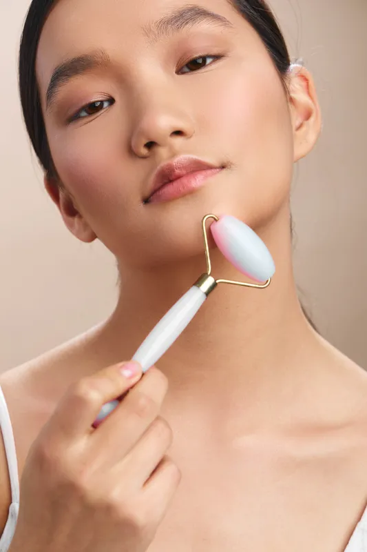 Asian woman uses a facial roller in a calm spa like setting, smiling softly as she treats her skin with gentle massage, creating a serene and rejuvenating beauty moment.