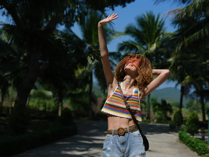 Smiling woman with curly hair wearing colorful top and sunglasses enjoys sunny tropical outdoors with palm trees and blue sky in summer.