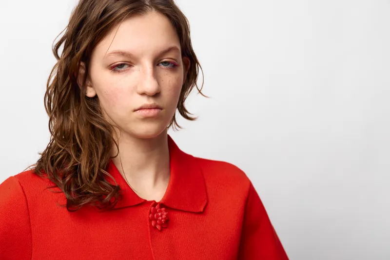 Young woman with wet hair and tired eyes wears red sweater, looking down with neutral expression on white background. Concept of fatigue, sadness, and emotional exhaustion.