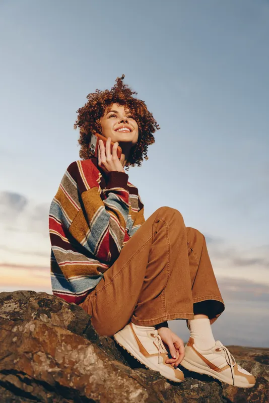 Woman lifestyle outdoors, curly hair woman in striped sweater and brown pants sits on a rock, hand on cheek, relaxed smile, scenic sky background, casual nature moment