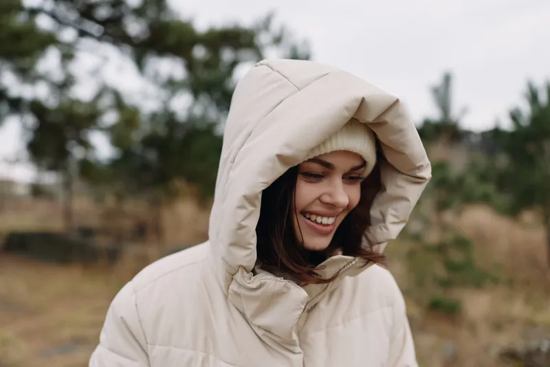 Hooded woman outdoors in a warm beige jacket smiles softly in a natural setting, capturing a casual portrait on a windy day with soft light and distant trees.