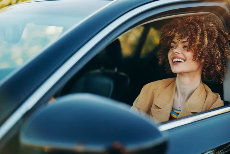 A smiling woman driver sits in a car, lifestyle shot with warm retro film tones, casual blazer, enjoying the ride. Concept for freedom, travel, and everyday joy.