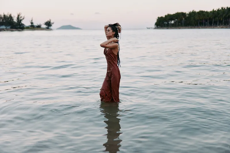 A solitary figure stands waist deep in calm sea water, wearing a rust dress and floral crown, captured in soft light with gentle ripples and a dreamy coastal mood.