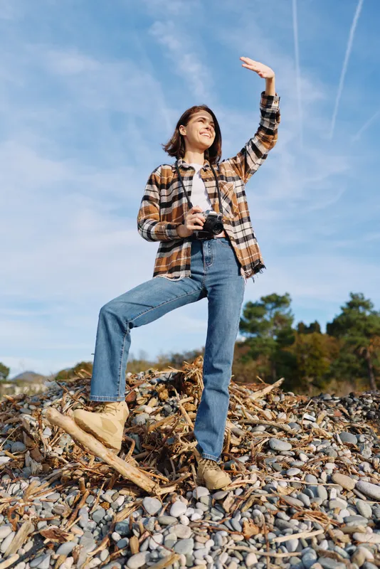 Outdoor woman with camera on rocky terrain under daylight sky, wearing plaid shirt and boots, posing with a raised arm for exploration and nature, a casual portrait capturing travel, adventure and