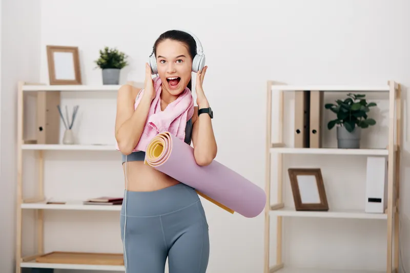 Fitness woman in athletic wear with a rolled yoga mat over her shoulder, headphones on, smiling in a bright home gym with shelves, plants and picture frames.