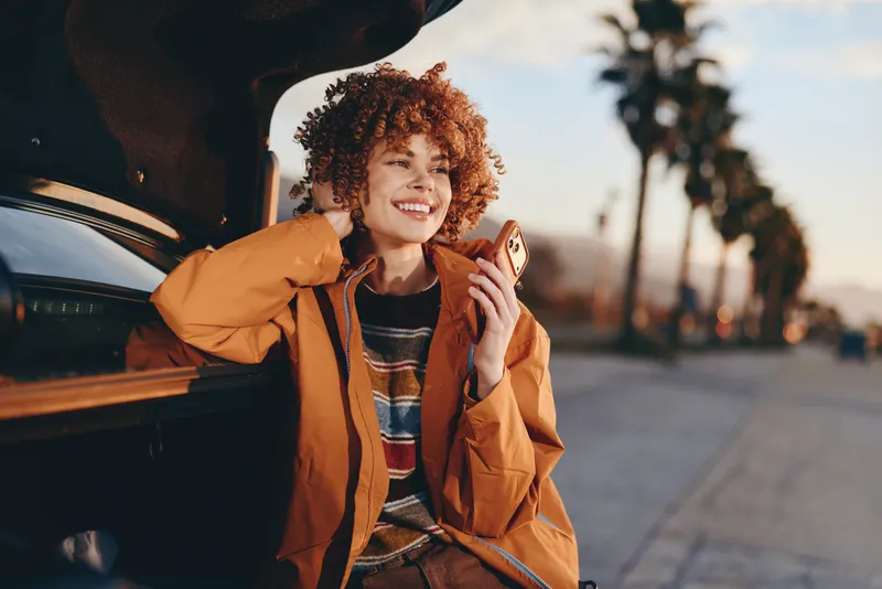 Woman lifestyle, curly hair, car, phone, travel outdoors, smile, orange jacket, standing beside open car door, holding smartphone, sunny street at sunset, warm light
