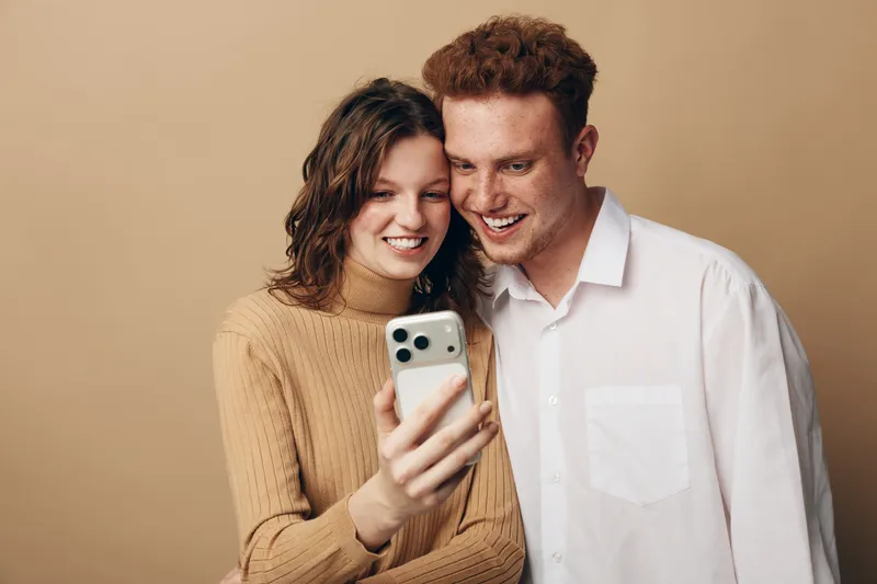 Happy young couple with curly hair smiling and looking at smartphone screen together. Casual clothing and warm beige background create friendly and joyful atmosphere.