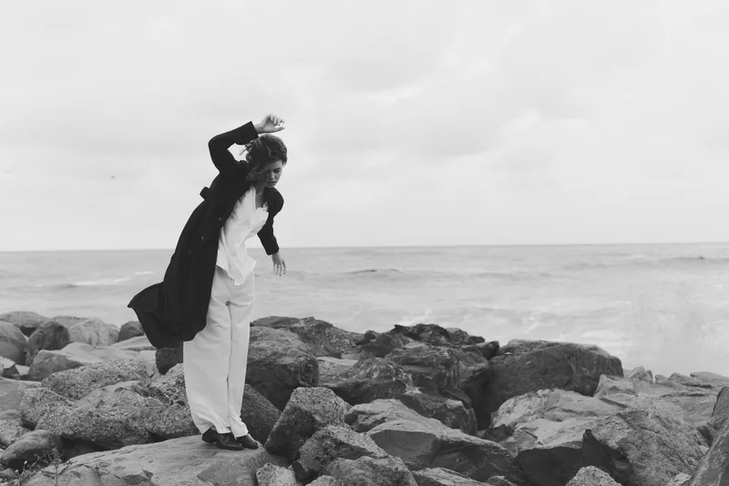 Monochrome scene of a lone figure on rugged rocks beside the sea, wearing a dark jacket and light pants, captured in a dynamic pose against a windy coastal backdrop