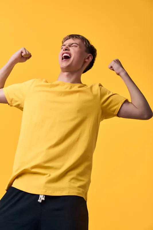 Celebration of joy and victory with a young man in a yellow shirt, arms raised in triumph against a matching backdrop, conveying happiness, energy and momentum.