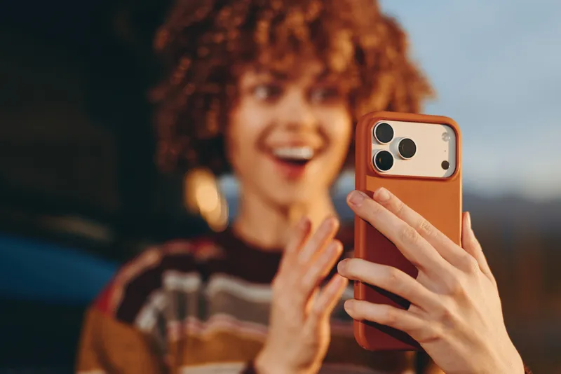 Woman lifestyle, curly hair smiling, holding a smartphone for a selfie outdoors, close up hands and phone, blurred background, warm natural light, casual mood during travel daytime