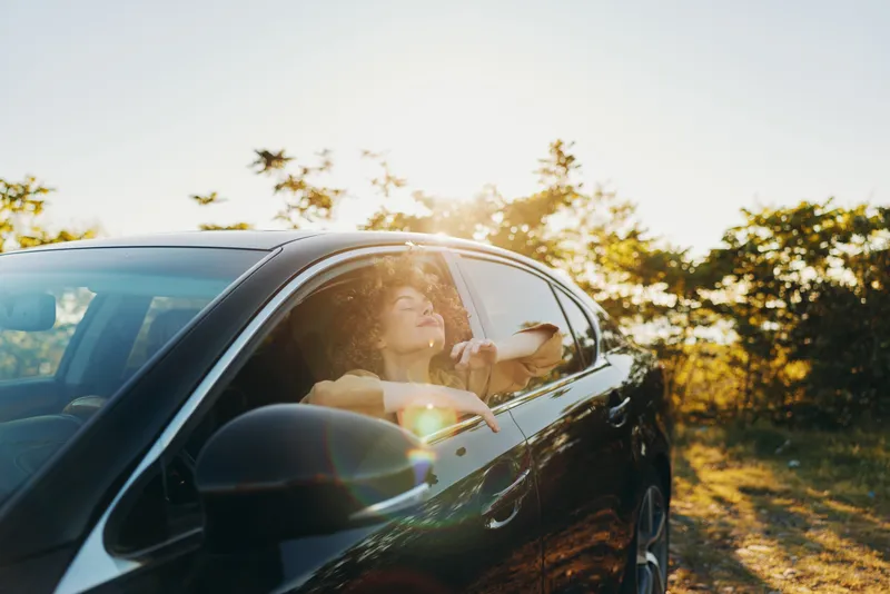 Young woman in a sleek car leans toward the sunlit window, smiling as warm retro film colors wash over the scene during a carefree daytime drive.