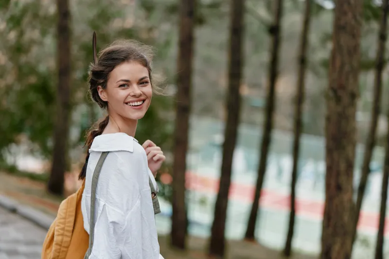 Young woman with a joyful expression wearing casual clothing and a backpack, smiling in a serene forest setting, embodying the essence of adventure and self discovery