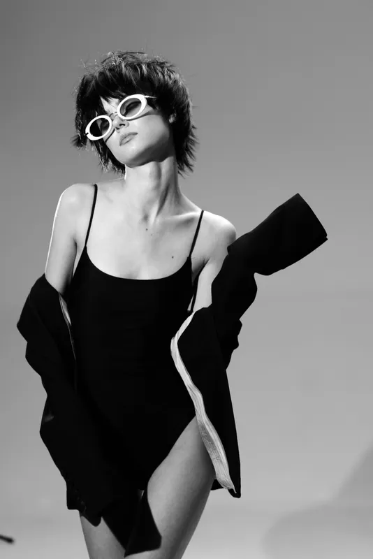 Black and white portrait of a stylish woman wearing sunglasses and a black swimsuit with a jacket on gray background.