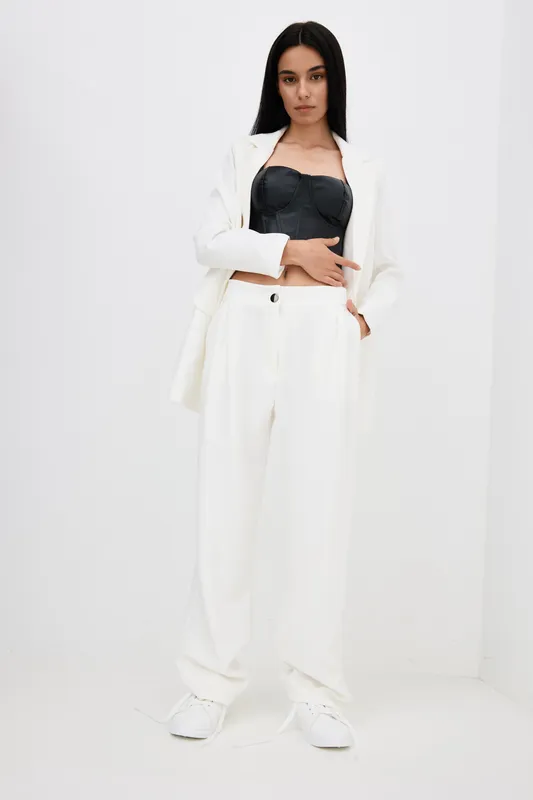 Young woman wearing a stylish white suit and black corset standing against a plain white background, showcasing modern fashion and minimalistic elegance.