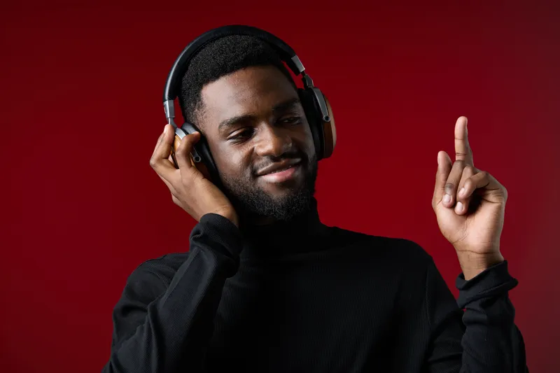 Young African man enjoying music with headphones, smiling against a red background, showcasing a vibrant and energetic mood that emphasizes relaxation and joy