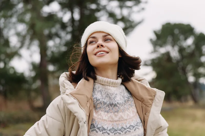 Woman in a light coat and knit sweater outdoors, looking up with a gentle smile as wind lifts her scarf, park setting and soft natural light creating a tranquil autumn scene.