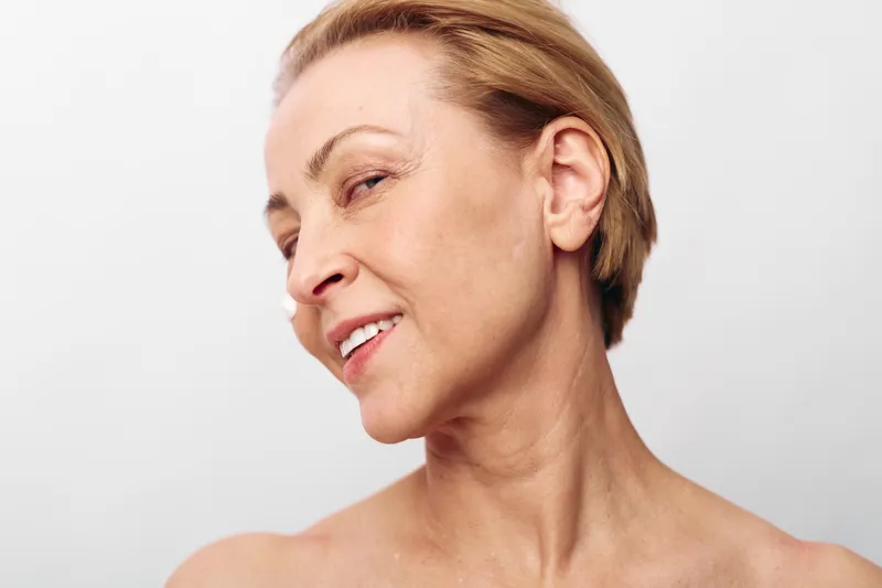 Close-up of confident mature woman with short hair showing natural skin texture and smile on white background. Concept for aging beauty, self-acceptance, and skincare routine.