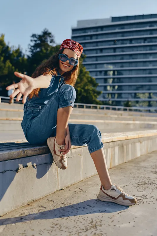 smiling woman in casual denim overalls and red bandana sitting outdoors on sunny day with sunglasses and relaxed confident pose
