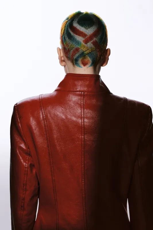 Woman with colorful geometric shaved hairstyle wearing a glossy red leather jacket, standing with her back against a white background in stylish fashion.