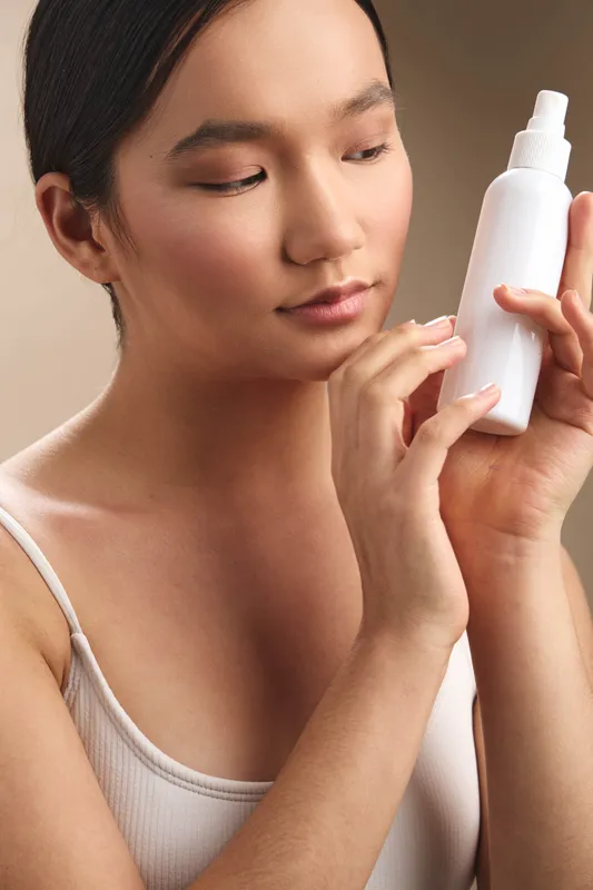 an asian woman studies a skincare bottle in a softly lit room, focusing on her routine. she looks calm and engaged, conveying a healthy approach to beauty and personal care.