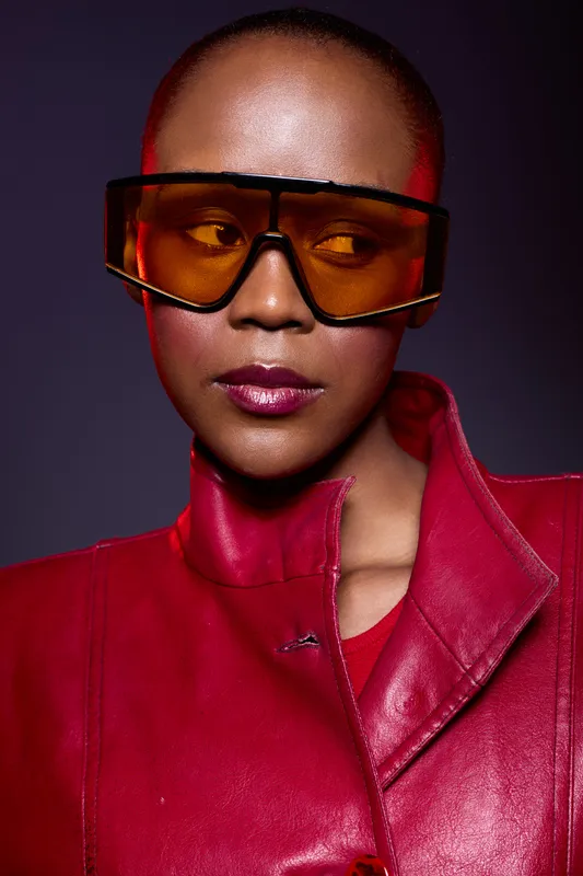 Close-up portrait of confident dark skinned African woman with shaved head wearing oversized orange sunglasses and red leather jacket. Studio lighting highlights stylish modern fashion look.