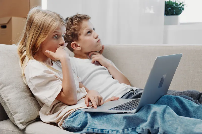 Two children sitting on a couch watching a laptop screen with thoughtful expressions in a bright living room. Concept of technology, childhood, curiosity, and learning.