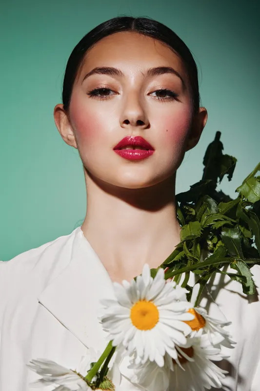 Close-up of a fashionable young woman with glossy red lips and long eyelashes holding a bouquet of white daisies. Soft teal background emphasizes modern beauty and creativity.