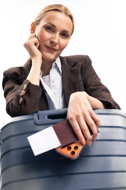 Young woman in brown jacket leans on large suitcase with passport and phone, smiling confidently. Concept of travel, tourism, vacation, and holiday preparation.