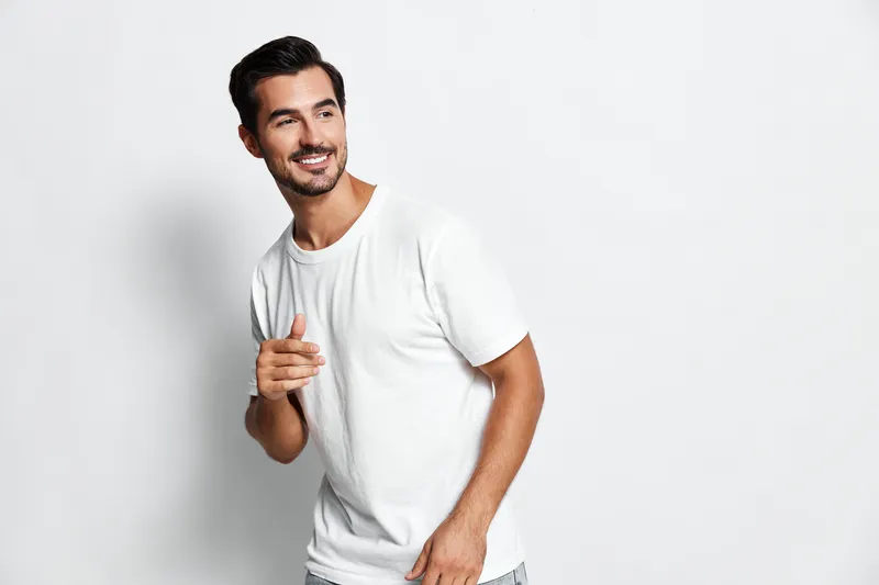 Smiling man in casual white t-shirt poses confidently against plain light gray background, radiating positivity and approachability. People lifestyle concept.