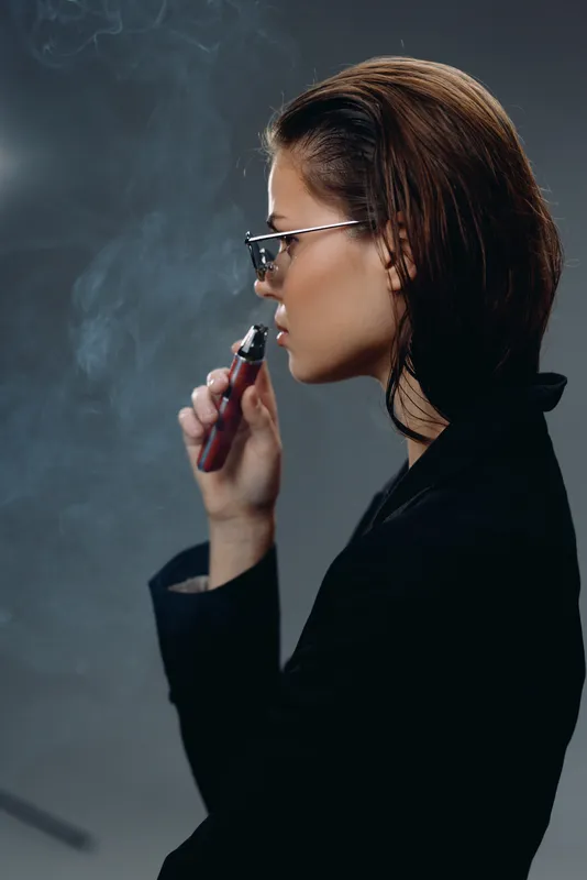 Profile of a young woman vaping with glasses, dark hair, and black clothing surrounded by vapor smoke.
