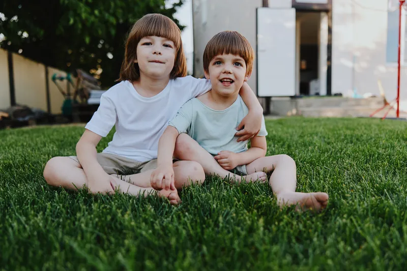 Two happy boys sitting on the grass, enjoying a sunny day outdoors. Their carefree smiles and playful poses reflect the joy of childhood friendship and playtime.