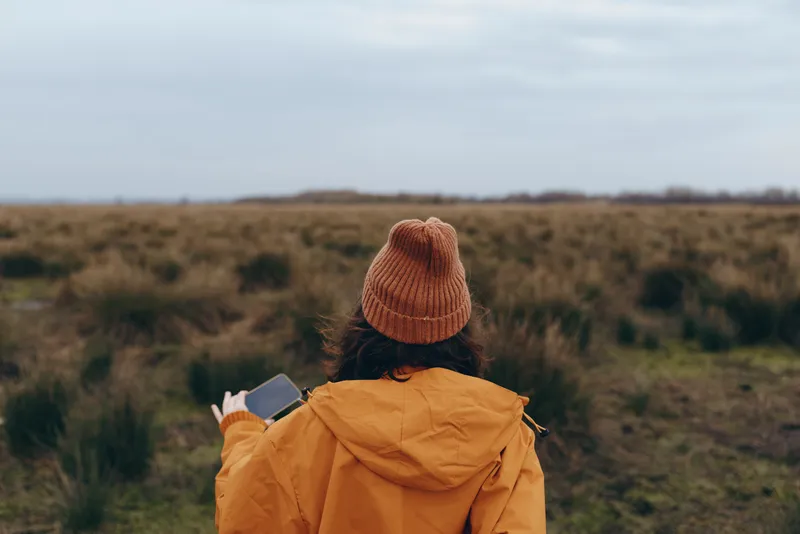 Woman outdoors in lifestyle moment, viewed from back, knit hat and yellow jacket, holding smartphone, open landscape, cloudy sky, calm rural atmosphere, quiet autumn fields, distant horizon