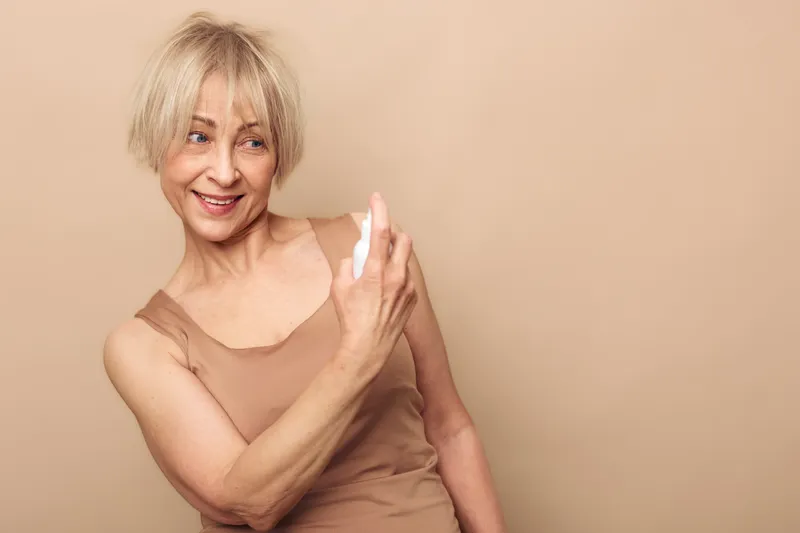 Mature woman with short blonde hair smiles while applying facial spray on beige background. Concept of skincare, hydration, self-care, and aging gracefully with beauty and wellness.